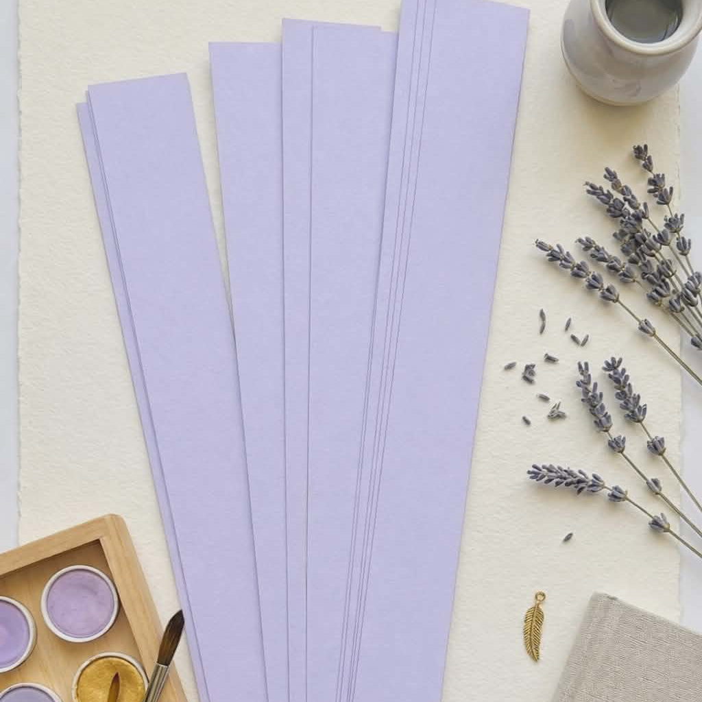 Lavender paper bead strips for rolling handmade paper beads, styled with watercolor paints and lavender flowers for jewelry making and craft projects.